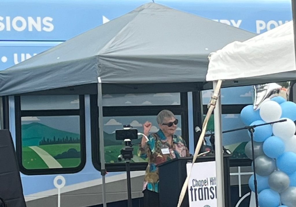 An older white woman speaking at a podium. Bus in background.