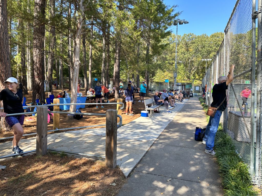 People standing around in a sitting area outside pickleball courts with trees in the background standing and sitting and looking at the pickleball courts