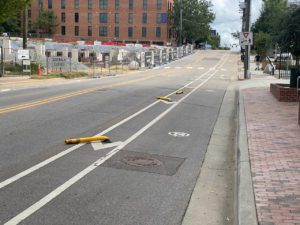 Picture of a two-lane road with a protected bike lane. The bike lane is buffered from the travel lane and there are flexi-posts in the buffer, but they have been knocked down.