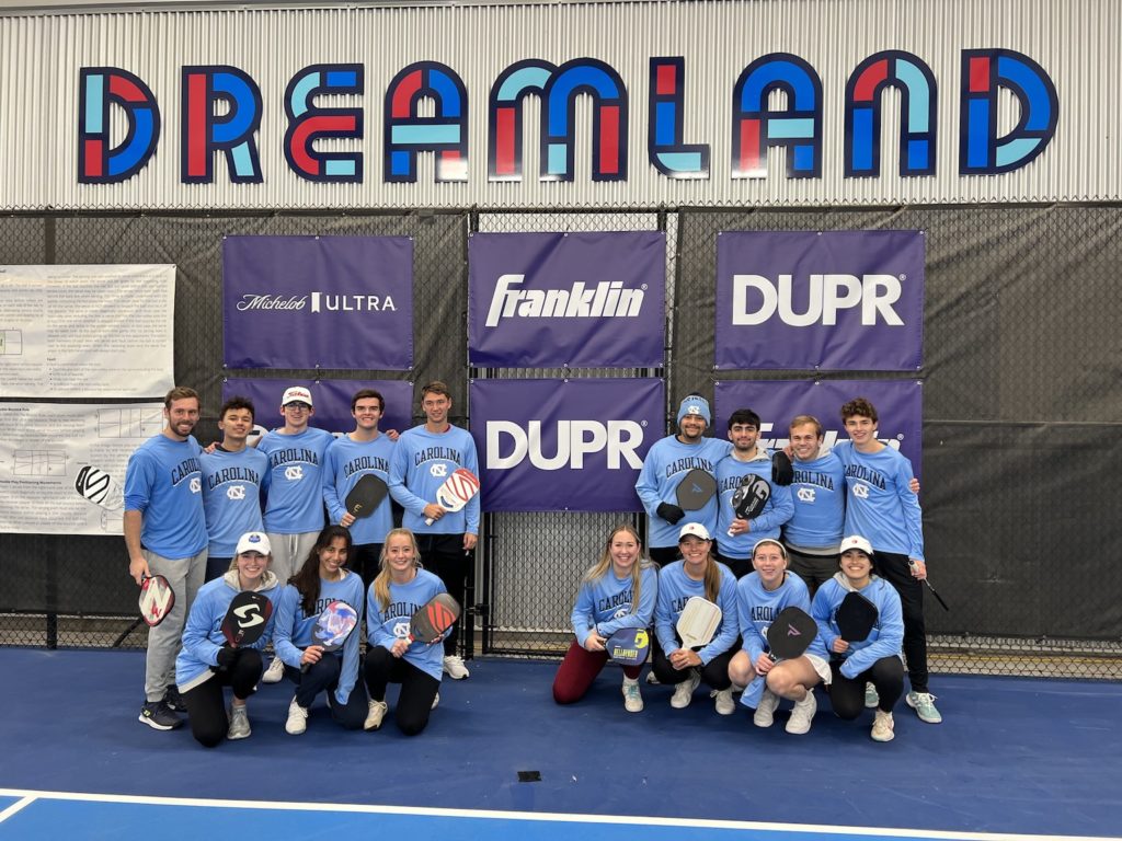 Photograph of the members of the UNC national championship pickleball team. There are 16 people pictured, in two groups of eight. On the left there are five men standing and thre women kneeling in front; each is holding a pickleball racket. On the right are four men standing and four women crouching in front, all holding pickleball rackets. The background is a fence with six purple banners with sponsor logos on them; two of the logos are covered by members of the pickleball team. At the top, in stylized colored text, is the word "DREAMLAND"