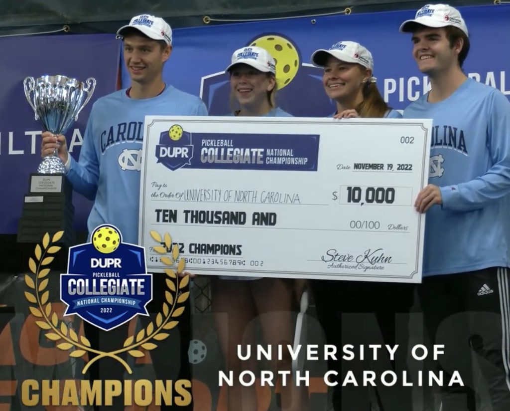 Photograph of four members of the UNC pickleball team, two men and two women. The man at the far left of the photo is holding a silver trophy. Collectively they are holding an oversized novelty check in the amount of $10,000 payable to the University of North Carolina. The bottom right of the photograph states in all-capital block white text, "University of North Carolina," and on the bottom left is the logo of the DUPR Pickleball Collegiate National Championship 2022, which has a logo with that text inside it inside a wreath with the word "Champions" below. 