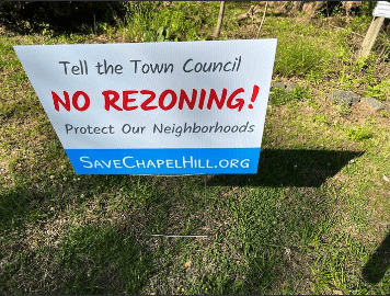 Photo of a yard sign on a grassy lawn that reads “Tell the Town Council NO REZONING! Protect Our Neighborhoods” in black and red text on a white background. At the bottom, on a blue background, is white text that states “SaveChapelHill.org”