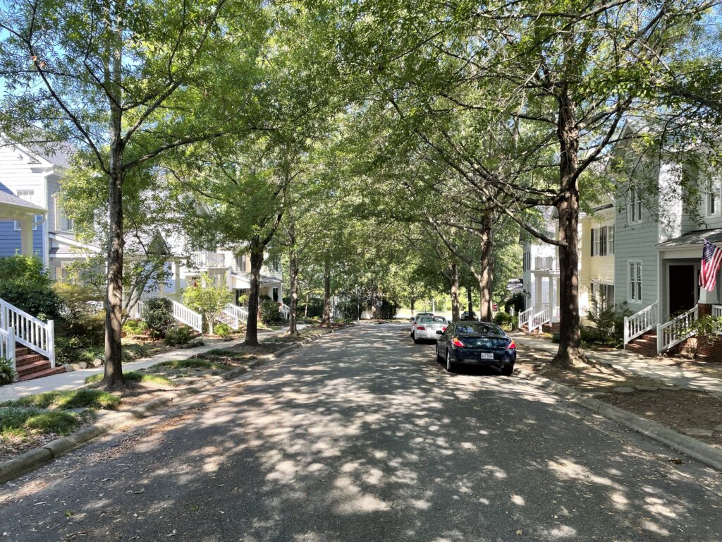 Photograph of a tree-lined street in Southern Village, taken during the daytime, with a lush tree canopy shading the entire street, a car parked on the right side of the road, and sidewalks and houses on either side