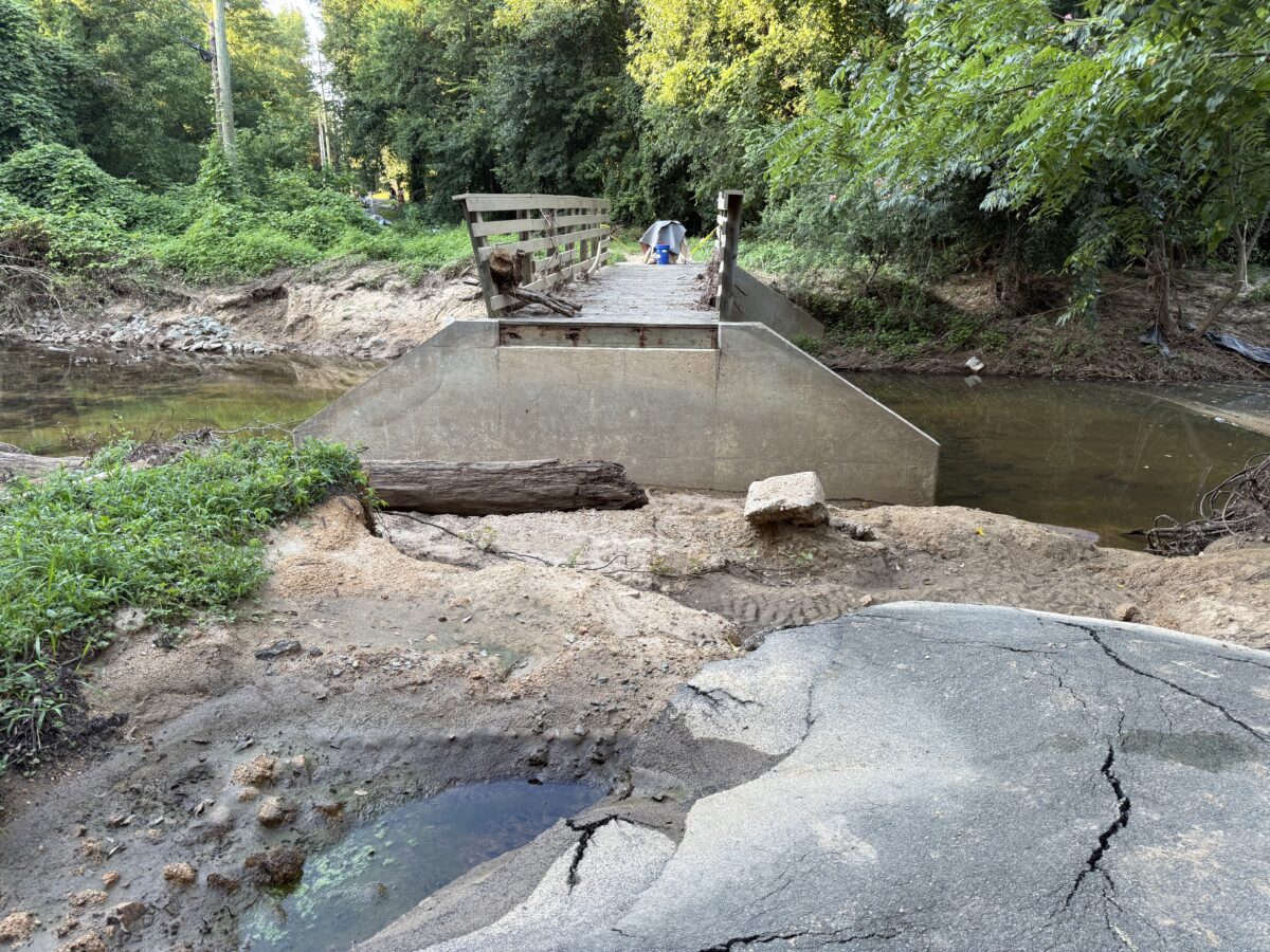 Photograph of a trail with broken asphalt and a large missing piece between the trail and a bridge over a stream