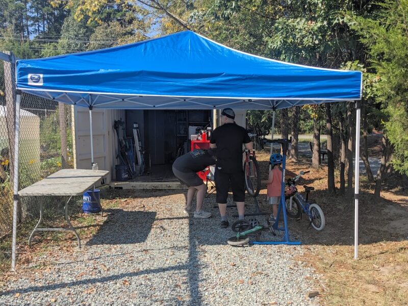 A mechanic works on a bike under a canopy while a child looks on