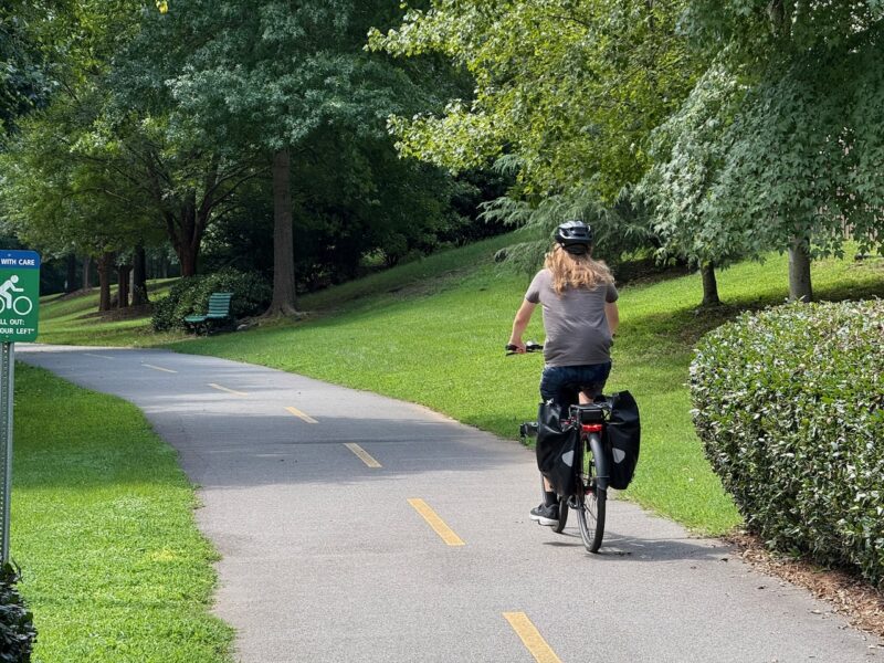 Photograph of a person biking on a paved trail. The person is wearing a gray shirt and black pants, and there are two panniers on the back rack of the bike. Off the trail to the side and front of the bicyclists are trees, grass, and a bench. Some trees cast shadows on the path..