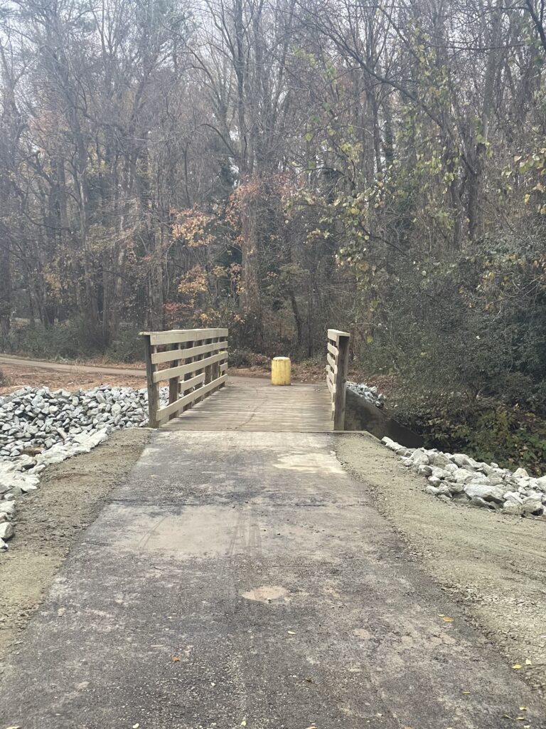A photos of a path along a trail leading to a bridge, with trees in the background.