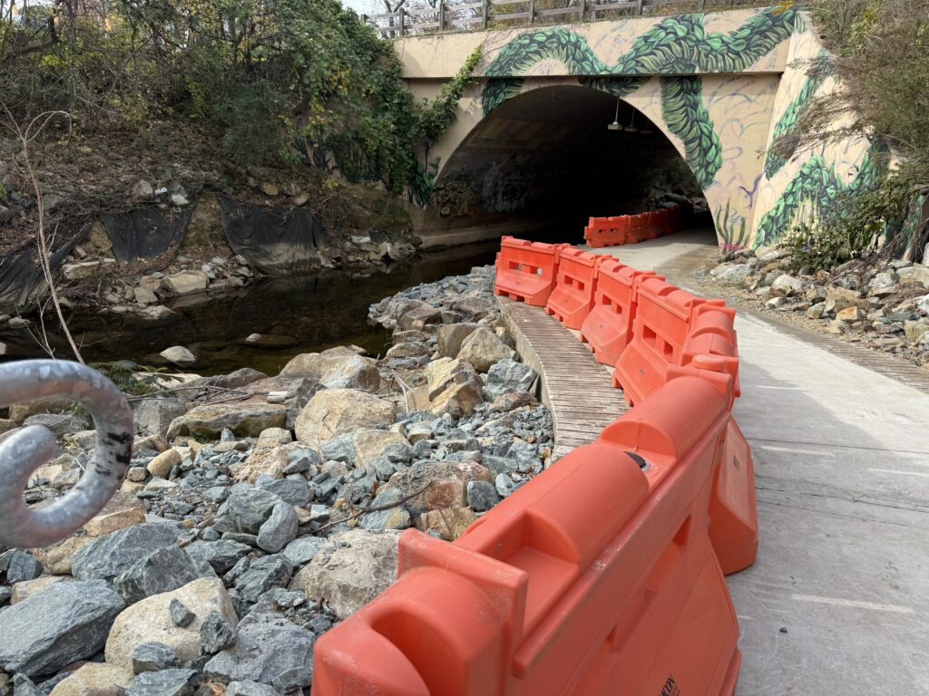 A photo of orange construction barriers along a trail. To the right is a concrete path; to the left are rocks piled up next to a creek with calm water. In the background is a bridge through which the creek and the train continue,.