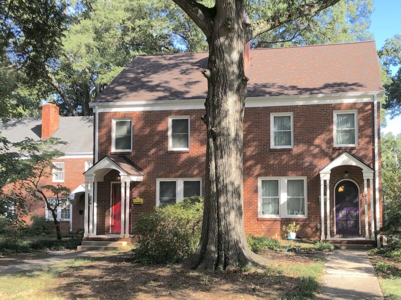 A photo of a red brick duplex house. It is setback on a grass lawn, with conrete paths leading to the front doors on either end of the house. The turnk of a very tall tree stands in between the two concrete paths in front of the house; The tree extends above the house and is so tall we don't see any leaves. The home is two stories tall, and there is a one-story portico in front of each front door.