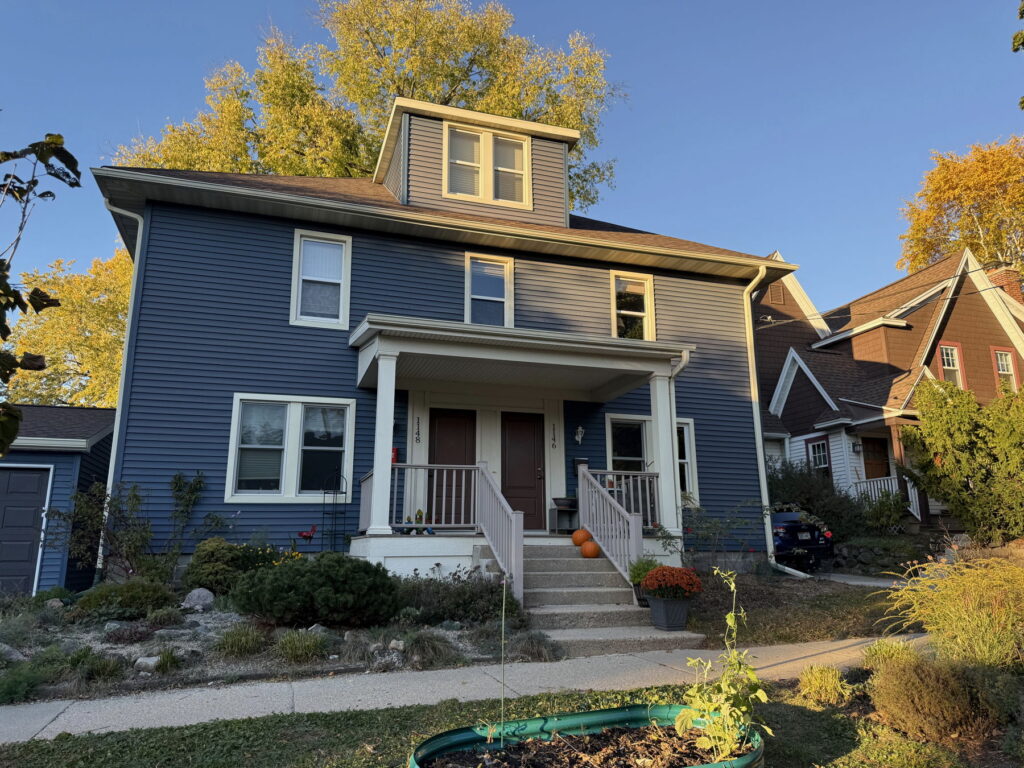 Photograph of a blue duplex house. It's painted blue, and seven steps lead up from the sidewalk to the front porch, where there are two side-by-side brown doors. The home on the left has the address numbers 1148 listed vertically to the left of the door, and the door on the right has the numbers 1146 listed vertically to the right of the door.