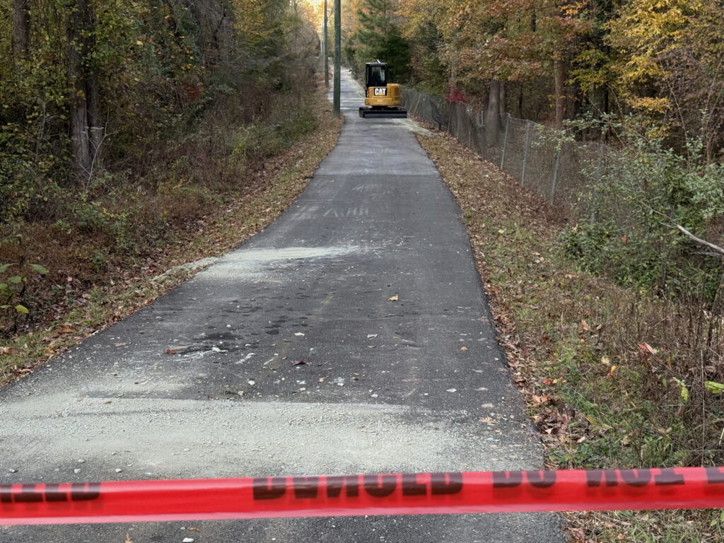 Photo of the Fordham Boulevard side path in Chapel Hill. On the bottom of the photo is red caution tape; above it is a paved asphalt path with fallen leaves on either side; on the left are trees, and on the right is a chain link fence with trees behind it. Near the top of the photo, down the path, is a large piece of machinery in yellow