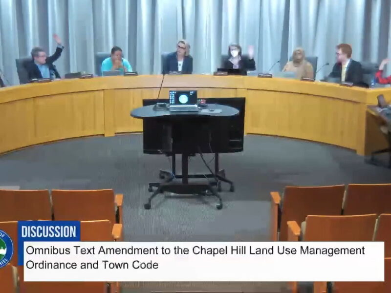Screen capture of the Chapel Hill Town Council on November 19, 2025, showing seven council members and the mayor sitting behind the Dias. Three council members have their hands up. Text on the screen at the bottom states: "Discussion: Omnibus Text Amendment to the Chapel Hill Land Use Management Ordinance and Town Code"