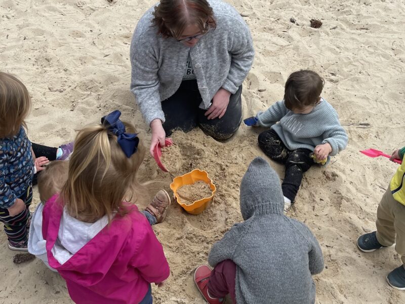 A picture of a pre-school teacher and five pre-school age children in a sandbox. No one's faces are visible