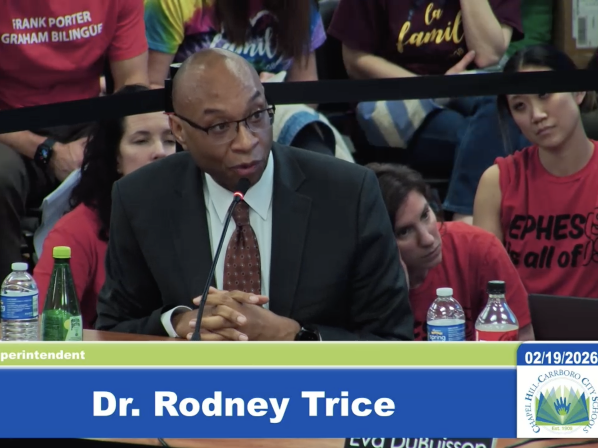 A man in a suit speaks at podium, with a crowd of students and parents behind him.